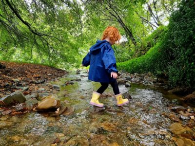 Child In Yellow Gum Boots And A Blue Coat Paddling In A Steam For Outdoor Play