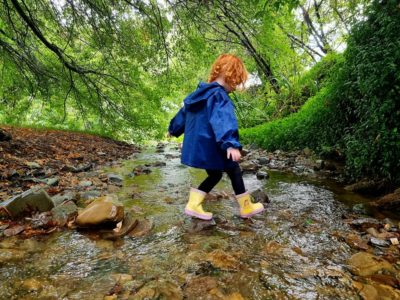 Child in yellow gum boots and a blue coat paddling in a steam for outdoor play