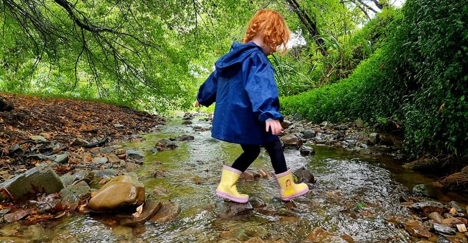 Child in yellow gum boots and a blue coat paddling in a steam for outdoor play