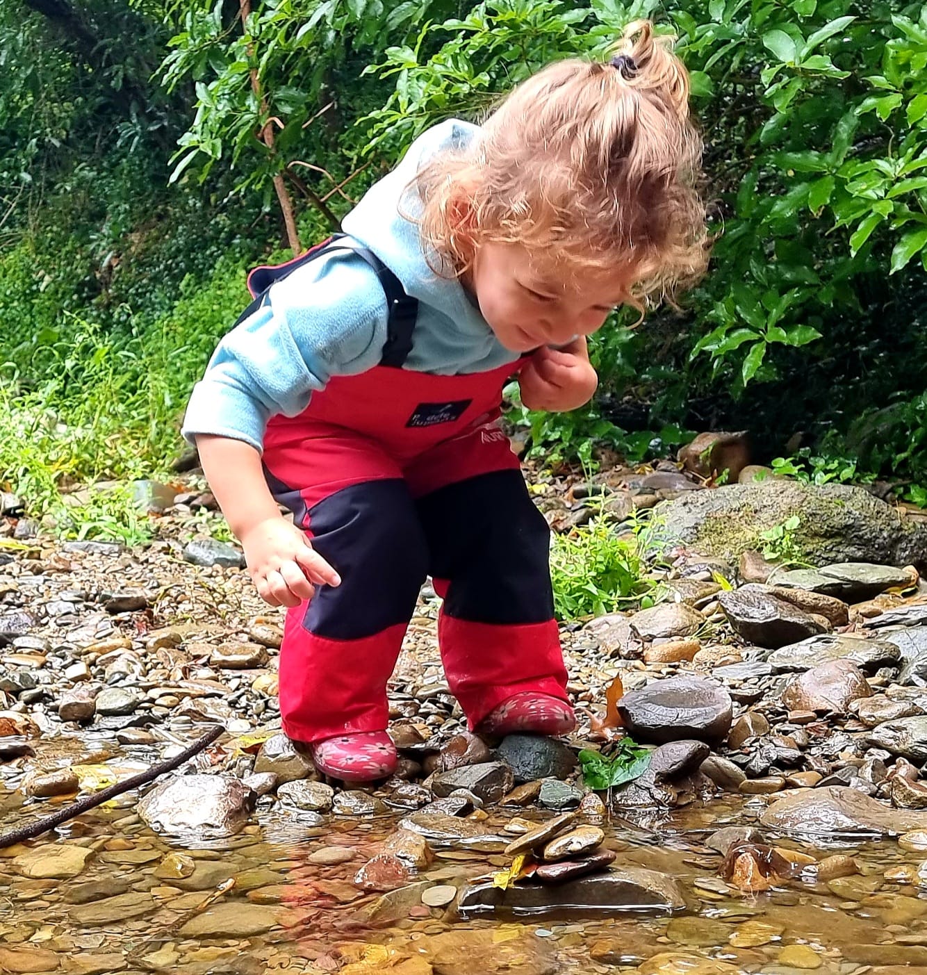 Child in red pants and gum boots stacking pebbles in a stream during outdoor play